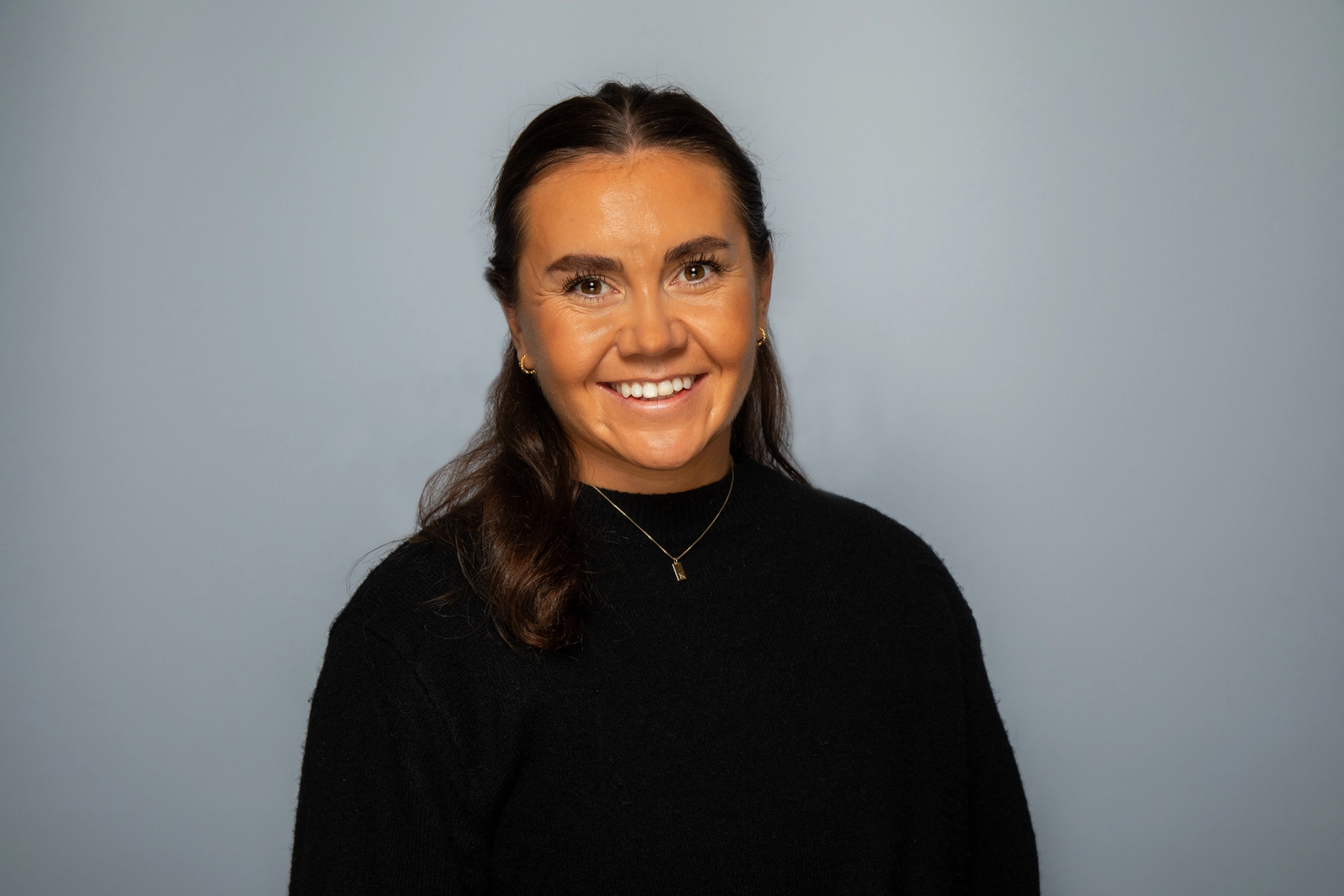 Dame med brunt, langt hår smiler mot kamera. Hun har på seg svart genser og smykke.