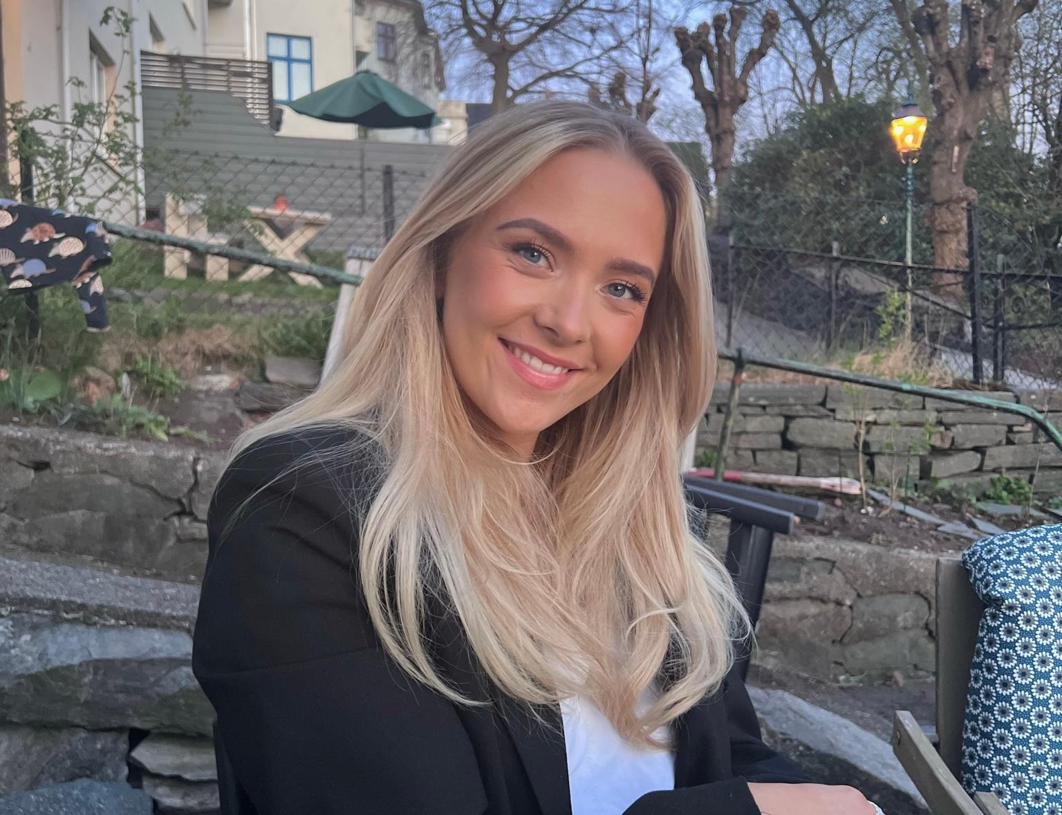 En ung kvinne med langt, blondt hår som smiler. Hun har på seg en svart genser og hvit topp, i et utendørs miljø med svakt kveldslys.