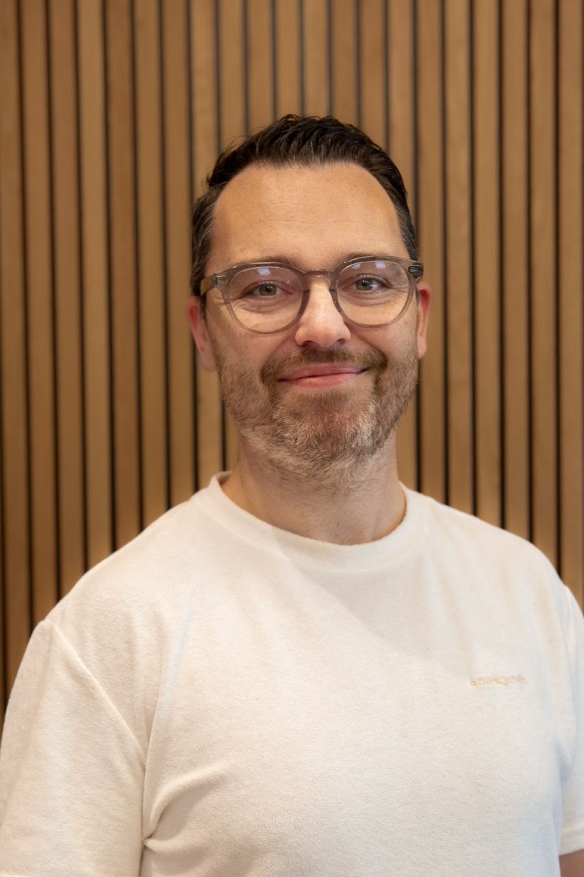 Portrett av en smilende mann med briller, kort mørkt hår og hvit t-skjorte framfor en vegg med trespiler.