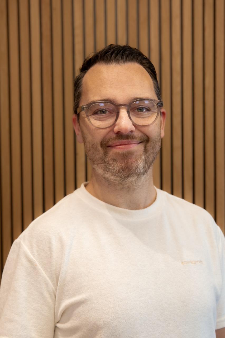 Portrett av en smilende mann med briller, kort mørkt hår og hvit t-skjorte framfor en vegg med trespiler.