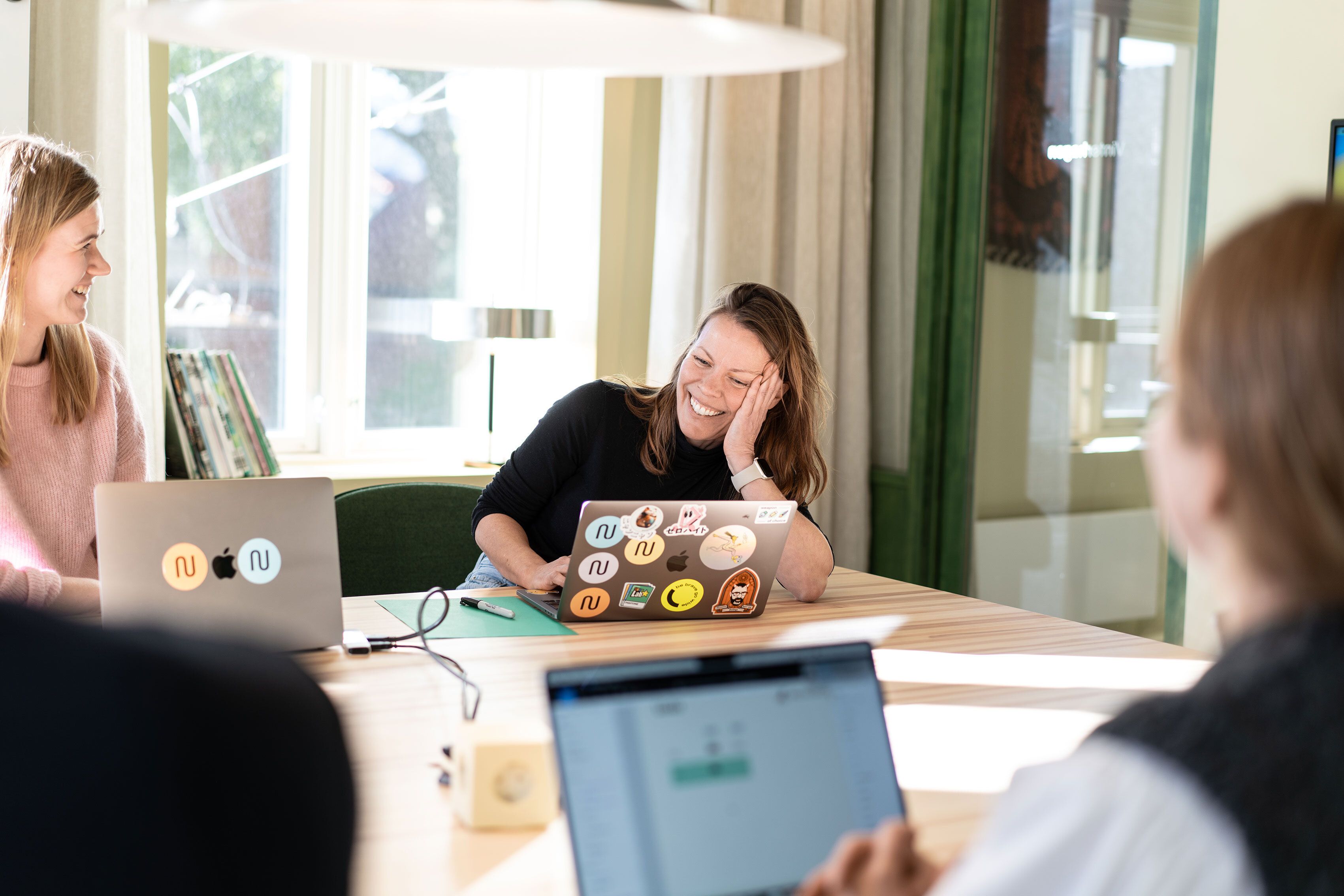 Smilende dame som sitter i kontorlandskap og jobber på laptop sammen med andre. 