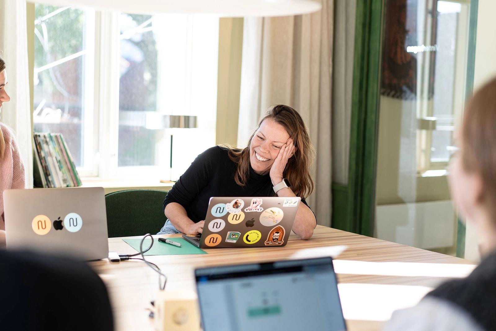 Smilende dame som sitter i kontorlandskap og jobber pĂĄ laptop sammen med andre.