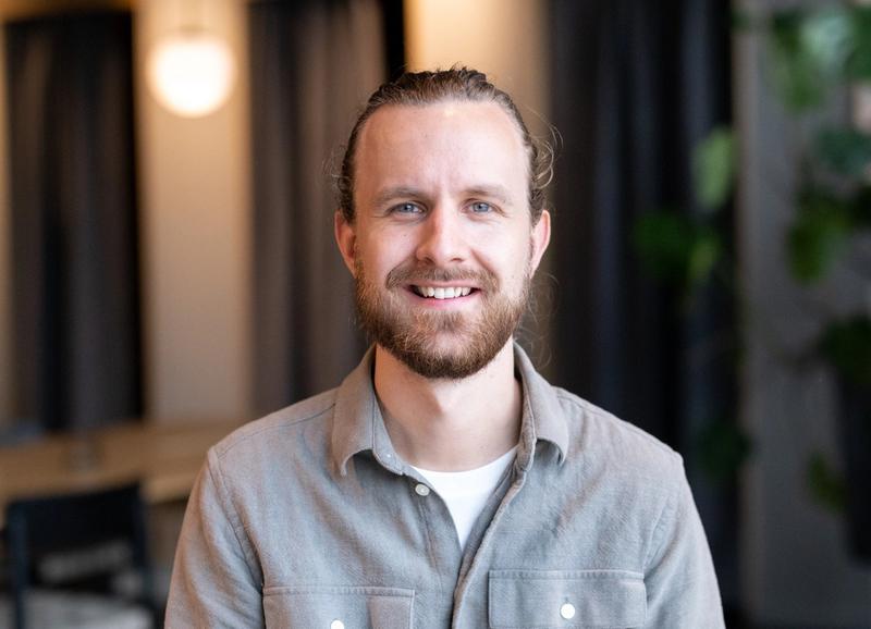 En smilende mann med skjegg og oppsatt hår i grå skjorte, i et kontorlokale med planter i bakgrunnen.