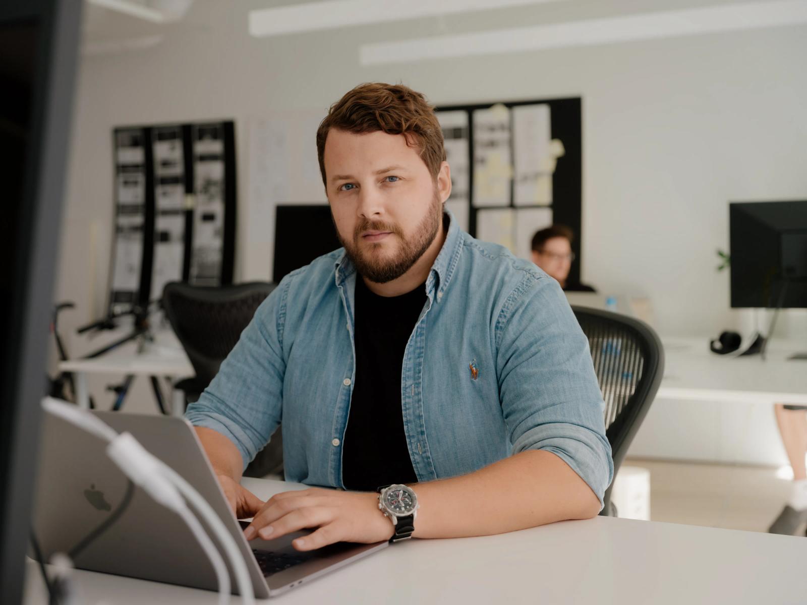 Mann med kort hår og skjegg sitter i hvit kontorlandskap og jobber på PC'en.