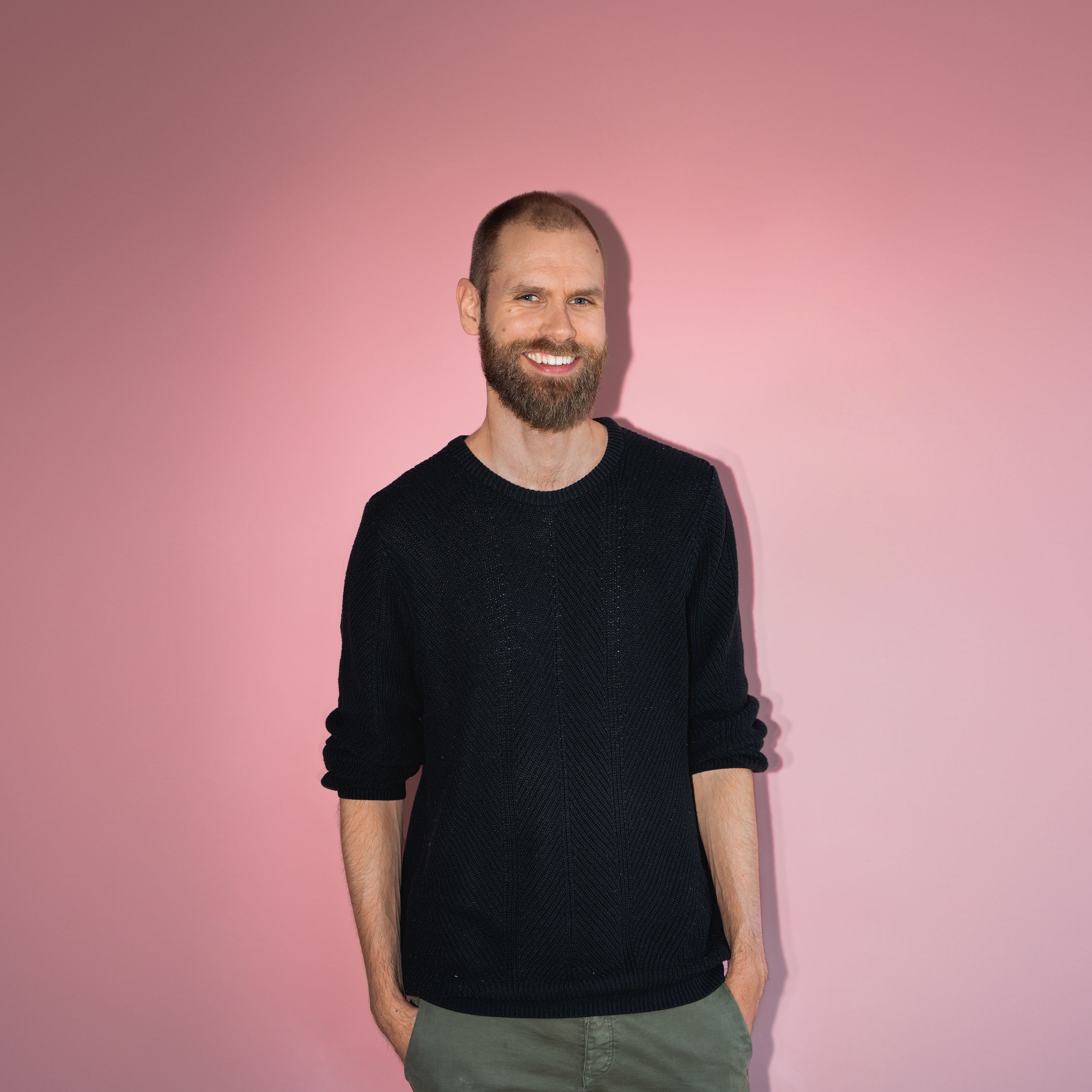 Mann med kort hår og skjegg smiler stort til kamera i studio med rosa bakgrunn.