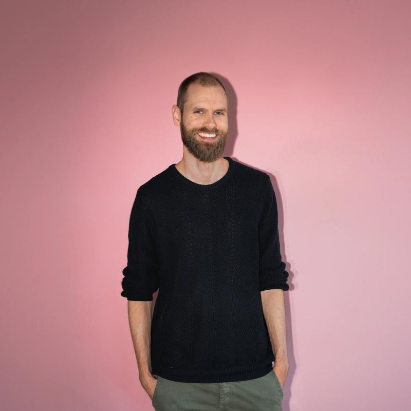 Mann med kort hår og skjegg smiler stort til kamera i studio med rosa bakgrunn.