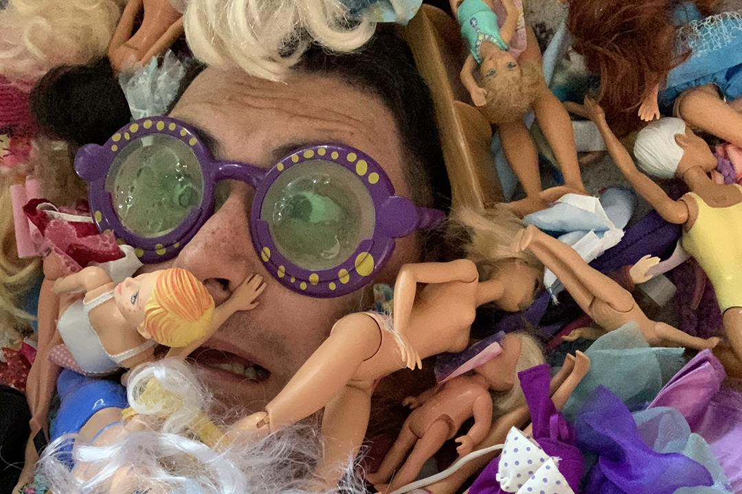 Adi har på store lilla solbriller og er dekket av Barbie-dukker