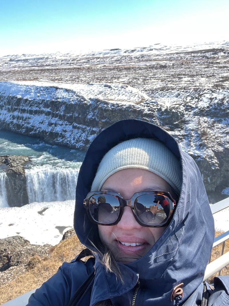 Selfie av Dame med vinterklær og solbriller. I bakgrunnen er det snølandskap og en foss. 