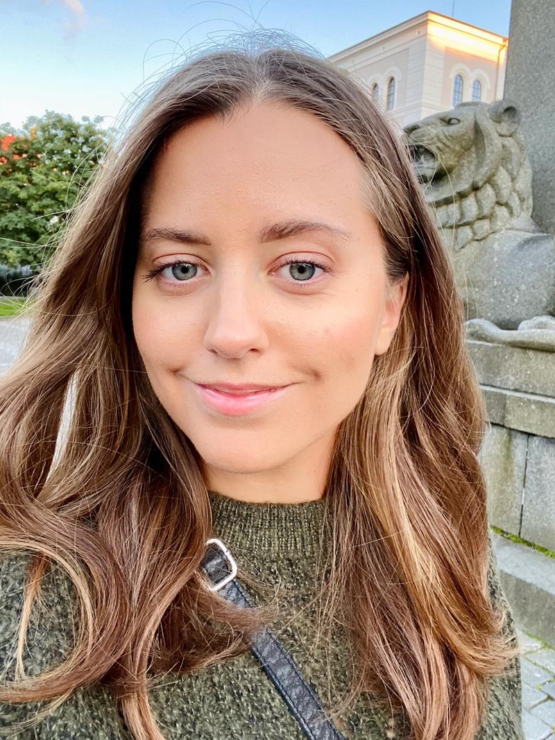 Bildet viser en selfie av Christine Heggum. Hun har lyst brunt hår, grønne øyne og har på seg en grønn genser. Hun står framfor løvestatuen som man finner utenfor Naturhistorisk Museum i Bergen.
