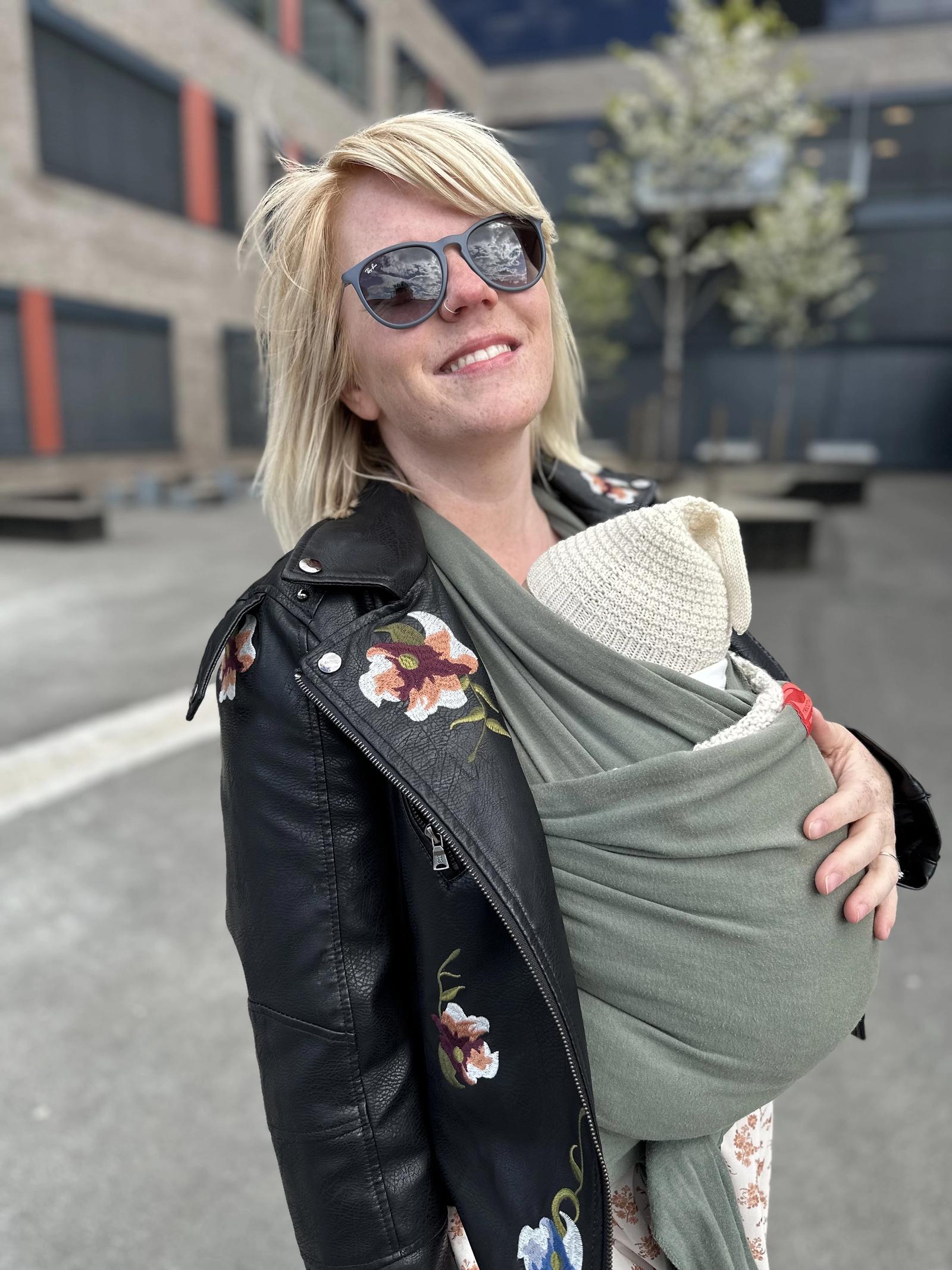 Susanne med solbriller på og med baby snurret rundt magen. Hun har blondt hår og en skinnjakke med blomsterdesign. 