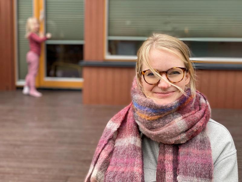 Dame med lyst hår og briller poserer utendørs med grå jakke og strikket skjerf i rosatoner.