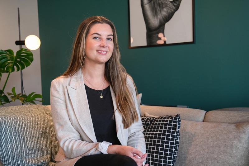 En dame med langt hår og grønne øyne sitter i en sofa. Rundt seg er det en plante, et bilde av en hånd og grønn vegg.