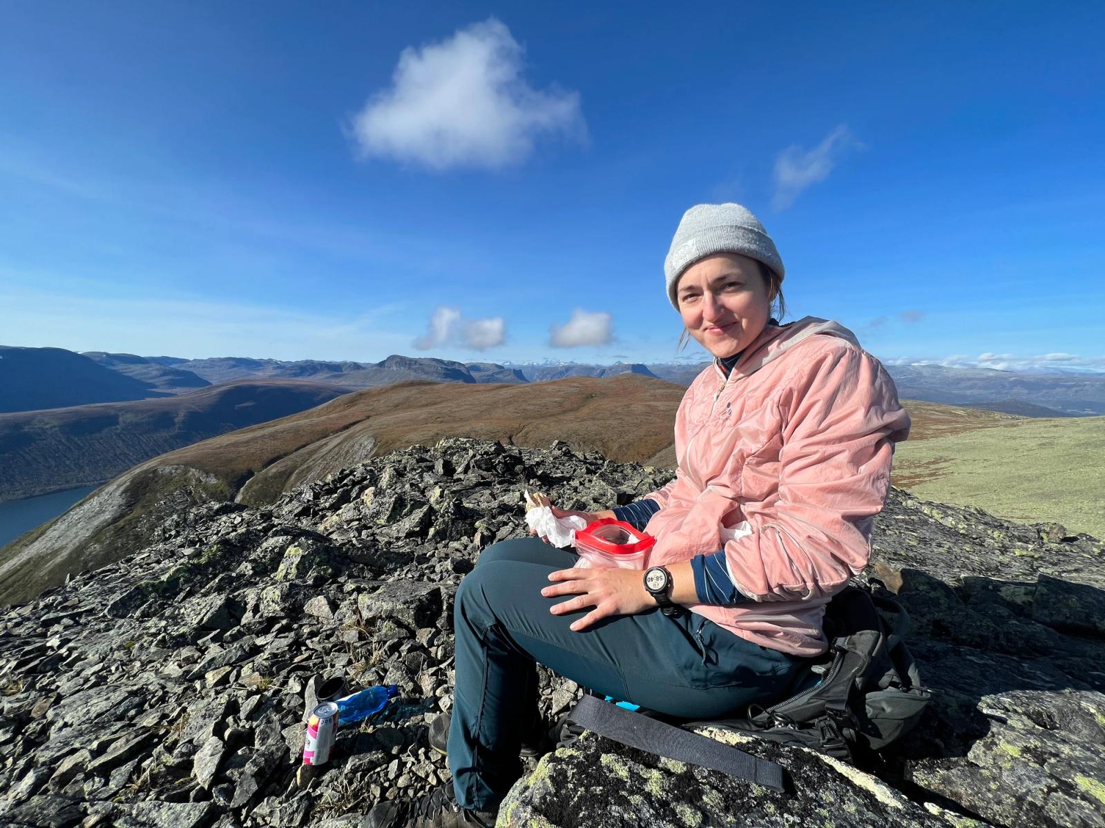 Dame i turklær sitter og spiser niste på fjelltopp.