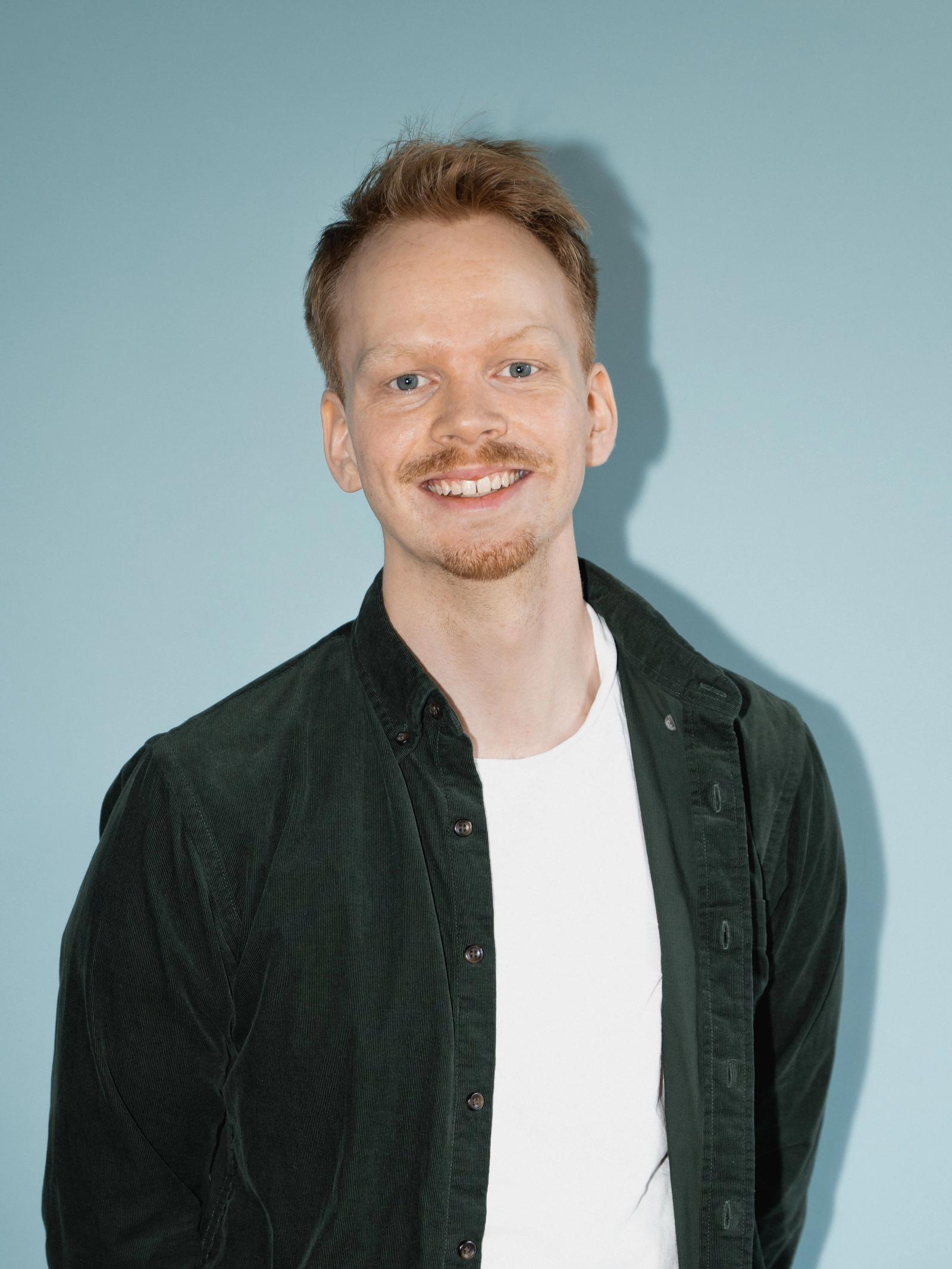 Mann med blå øyne, kort hår og bart står inntil en blå vegg og smiler
