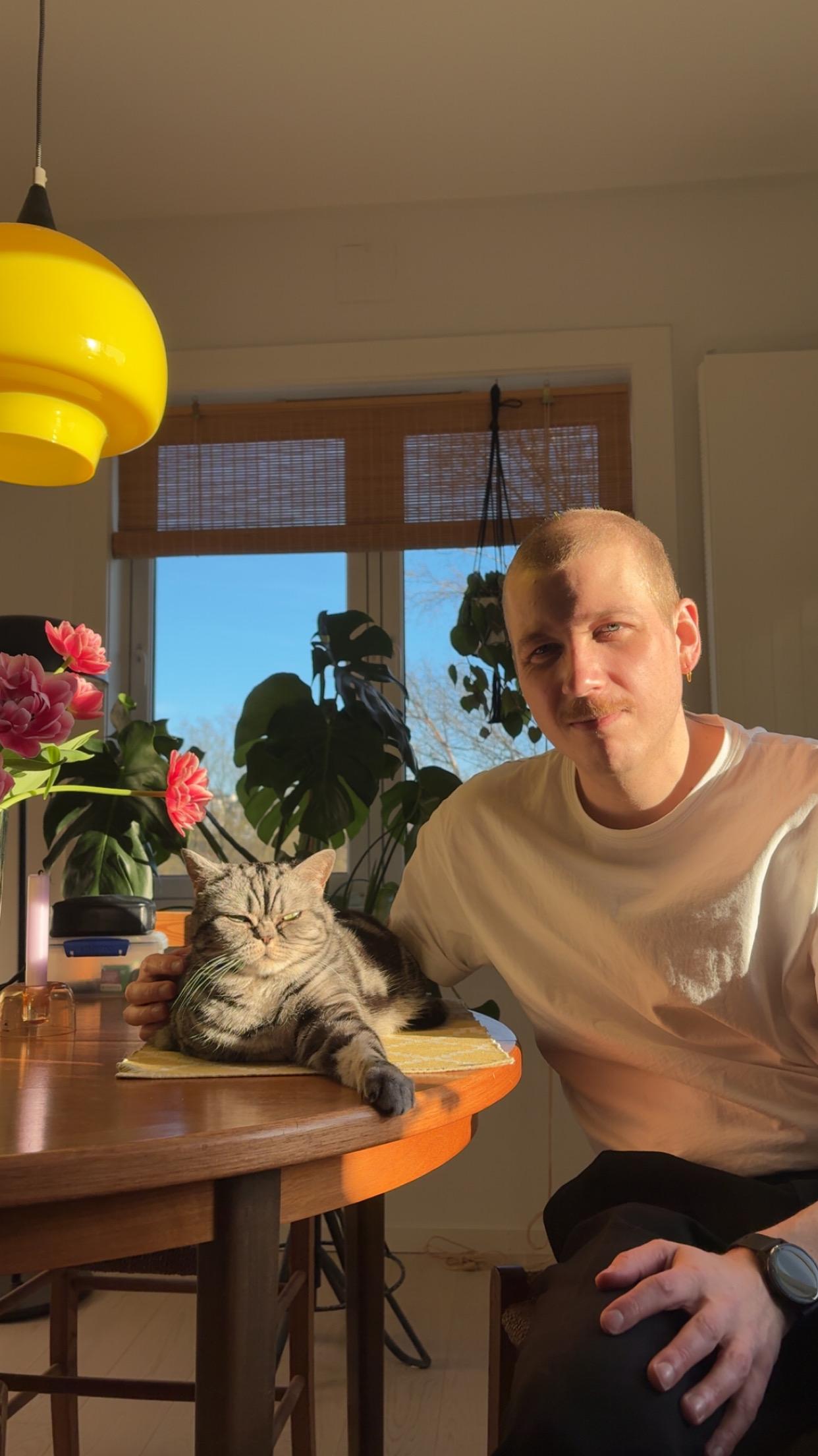 Mann sitter ved et kjøkkenbord i sollys med hånden på en gråstripet katt. Det er blomster og planter i vinduet bak.