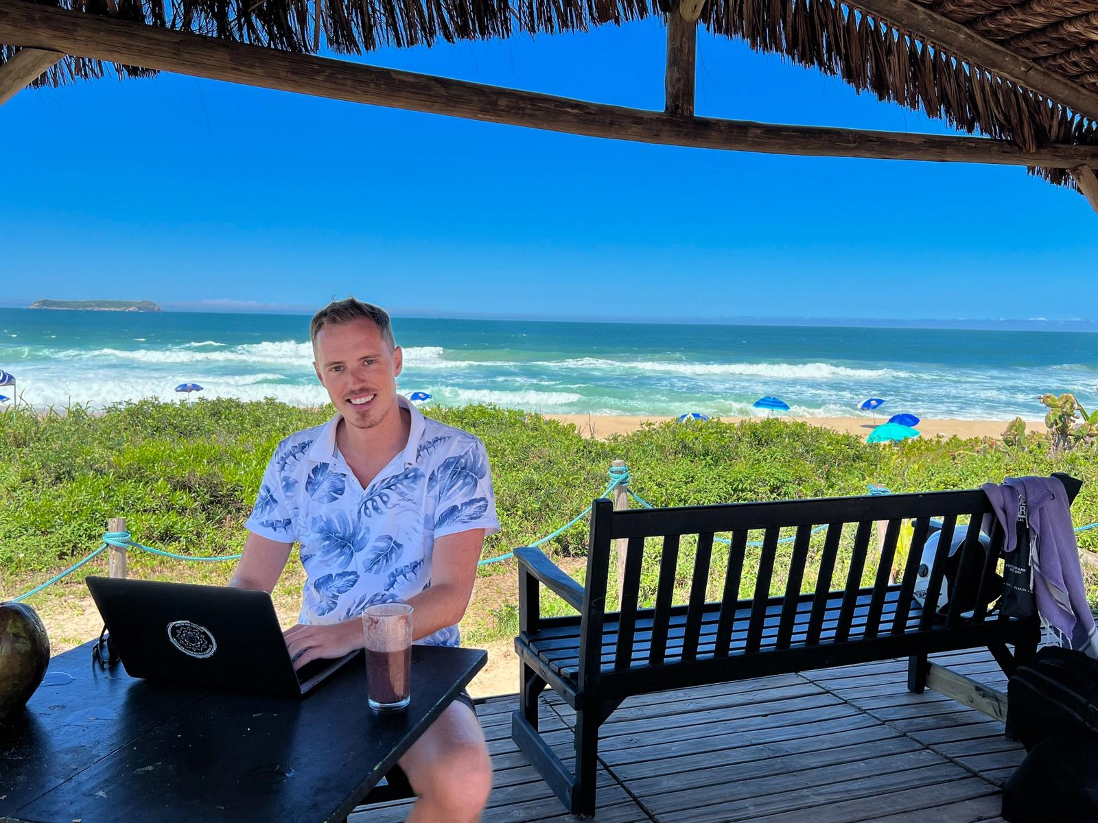Eirik jobber på laptopen på balkongen med strand, sjø og blå himmel bak seg.