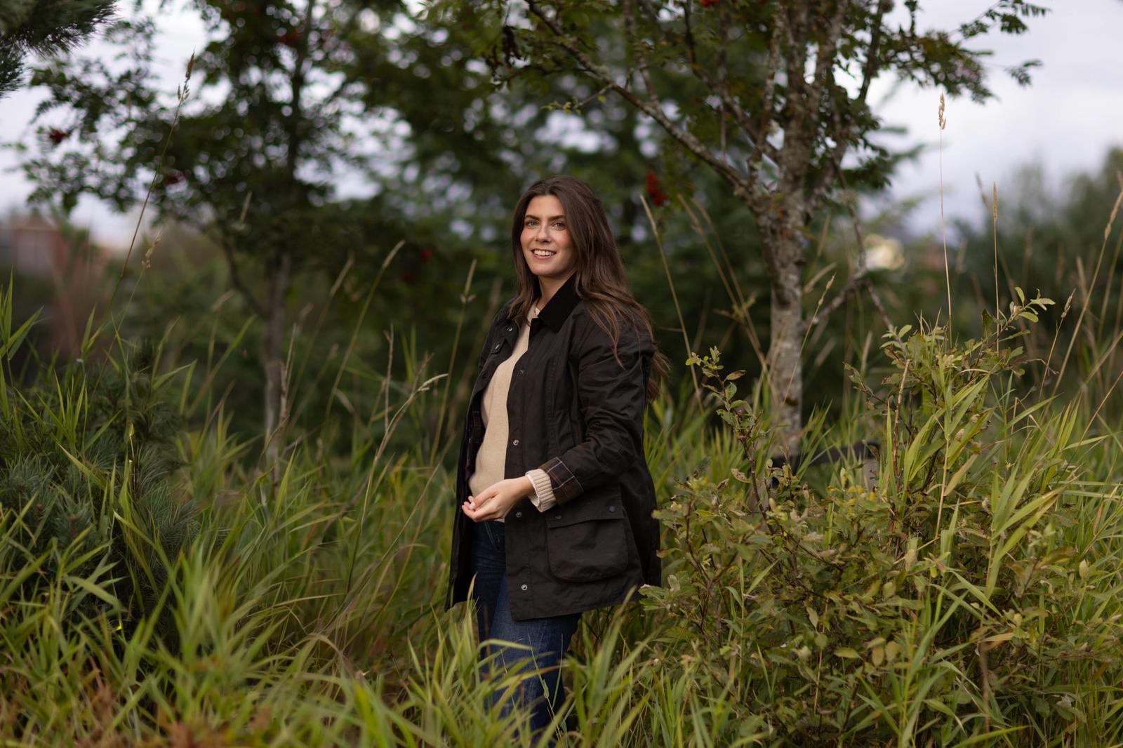 Dame med beige genser og svart jakke står i høyt gress i skogen og poserer for kamera.