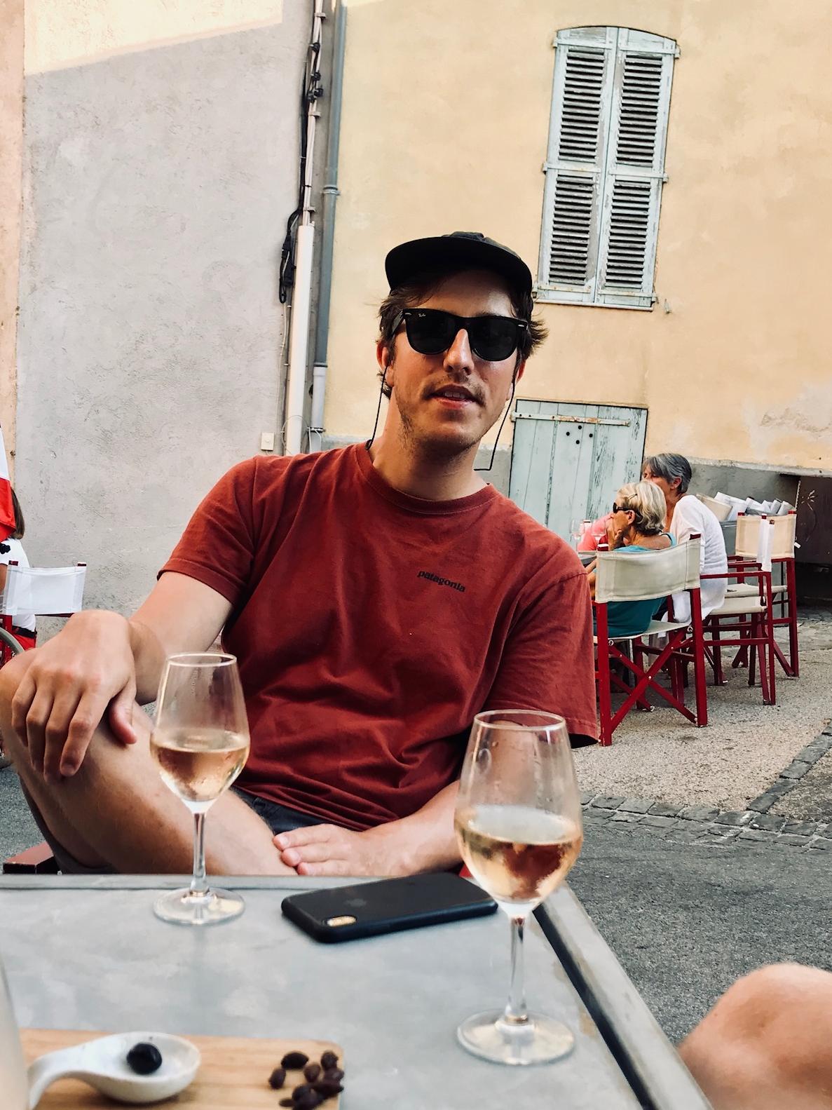 Mann med solbriller og caps sitter utendørs i bakhage og drikker rosévin og spiser på oliven.