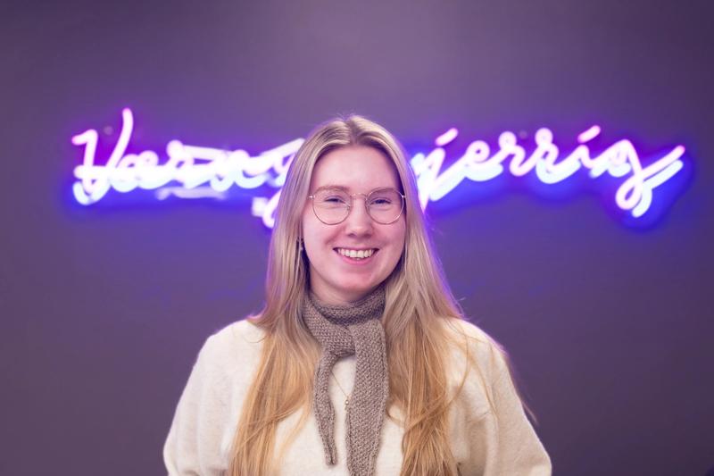 Dame med lyst, langt hår og biller med neon-skilt i bakgrunnen.