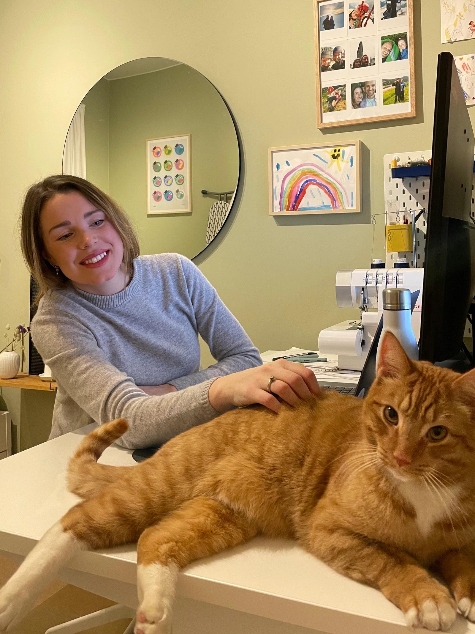 Dame sitter med PC-skjerm og klapper på en orange katt som ligger på skrivebordet.