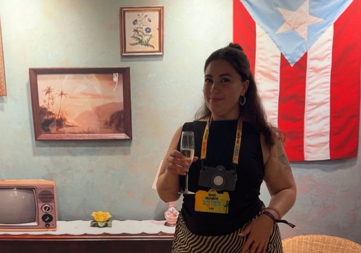 Dame med champagneglass og papirkamera rundt halsen står i et kunstig hjem med Puerto Rico flag i bakgrunnen.