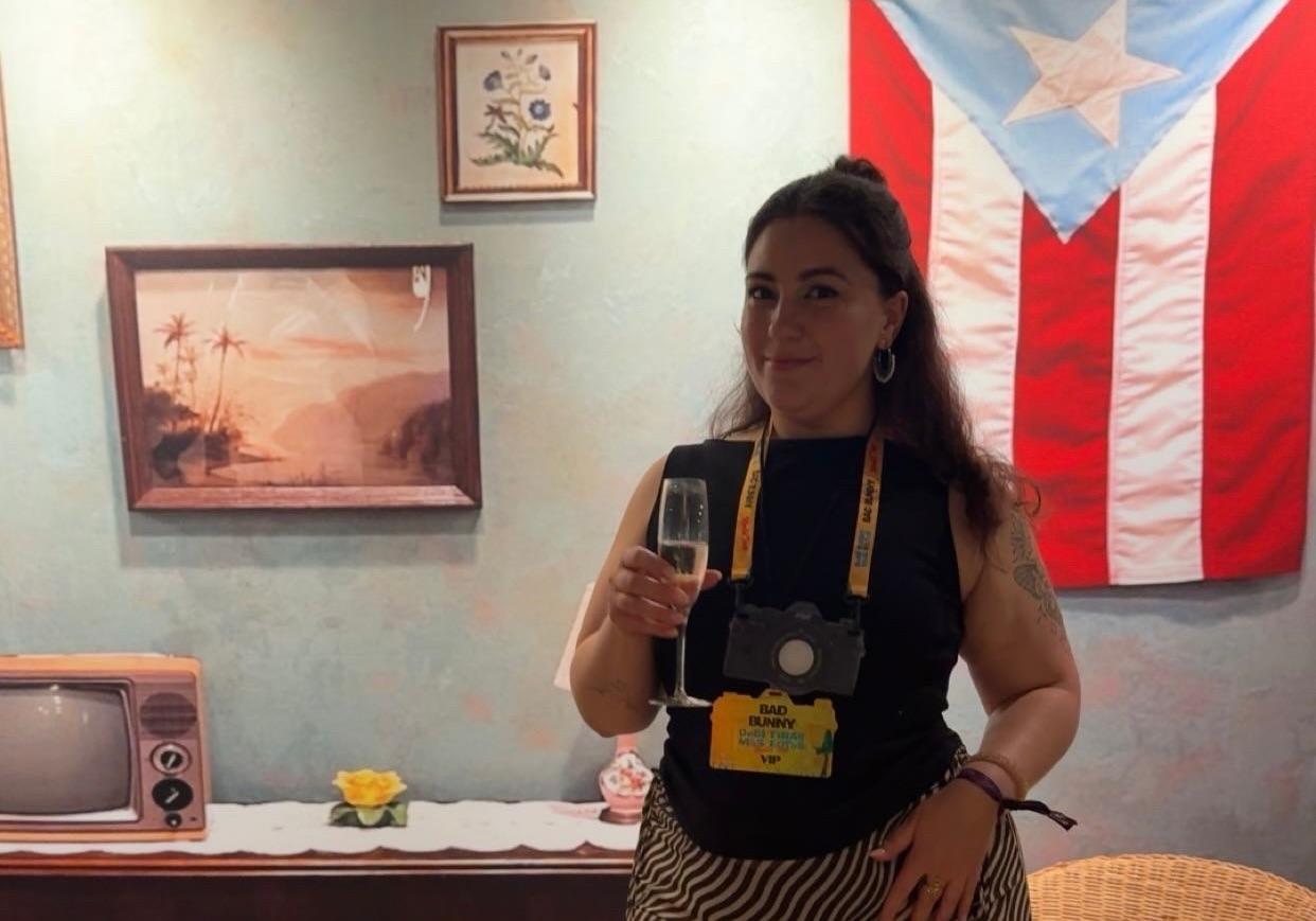 Dame med champagneglass og papirkamera rundt halsen står i et kunstig hjem med Puerto Rico flag i bakgrunnen.