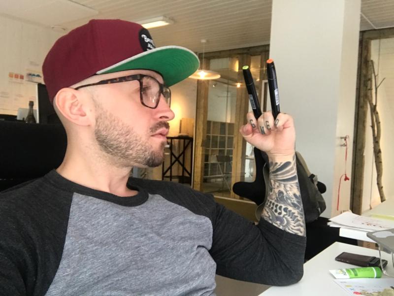 Mann med caps, briller og tatoveringer på armen og fingrene. Han har to tusjer mellom fingrene.