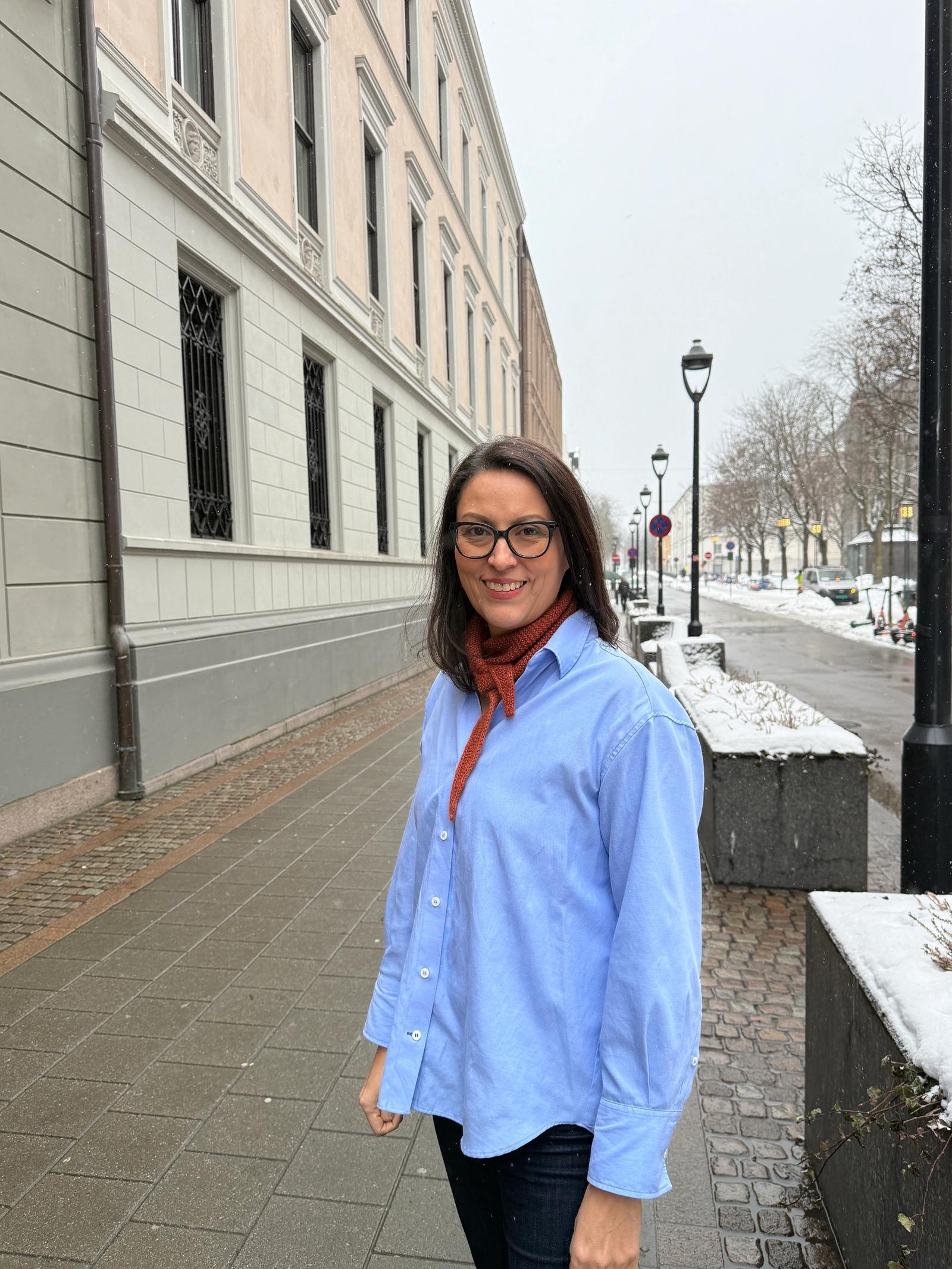 Kvinne med blå skjorte, mørkt hår og brilelr står utendørs.