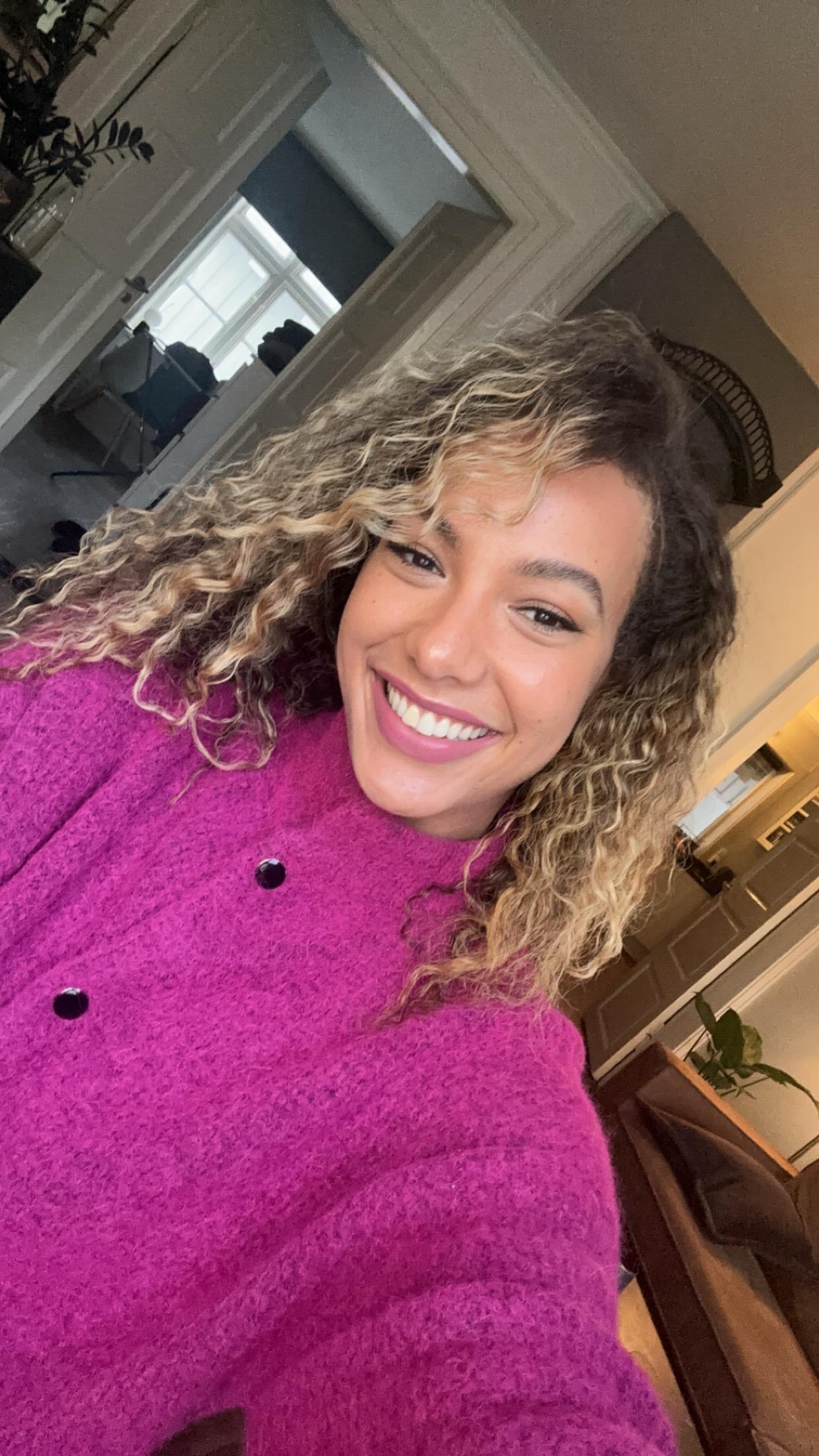 En dame tar selfie hjemme. Hun har på seg knall rosa genser, har krøllete hår med lyse tupper, brune øyne og et stort smil.