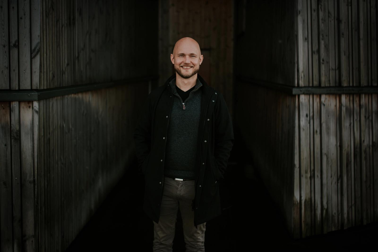 Erling står med en mørk bakgrunn, i en frakk og smiler