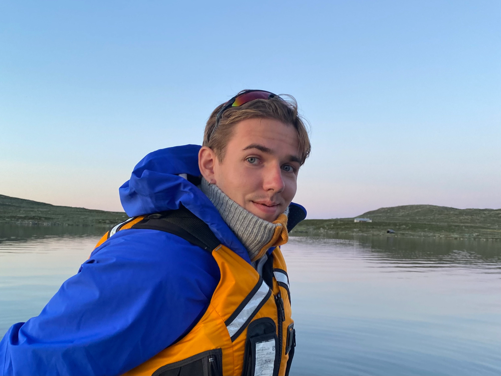 Mann med blå jakke og redningsvest sitter med et stille vann på fjellet i bakgrunnen.