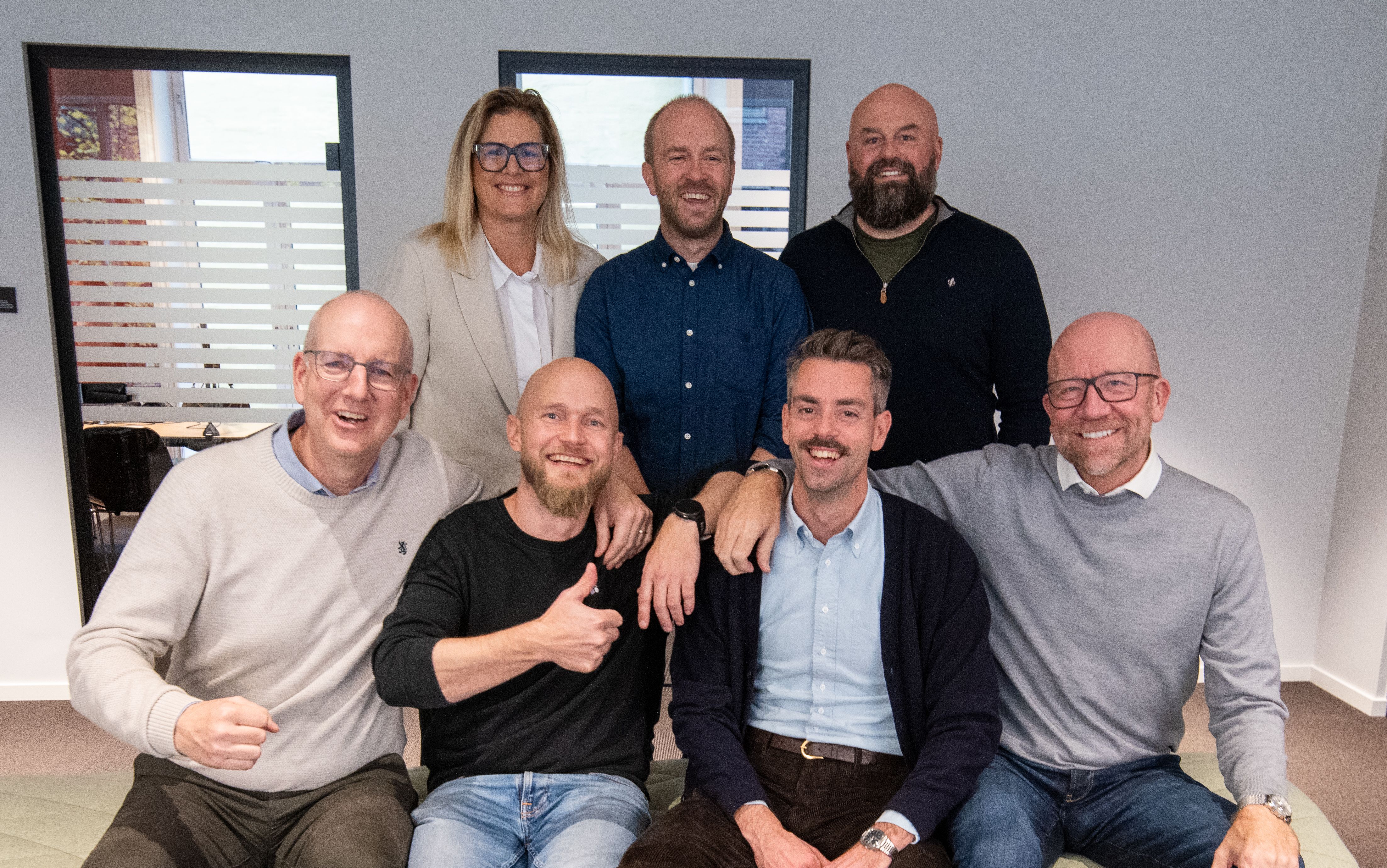 Seks menn og en kvinne sitter og står inne på et kontor