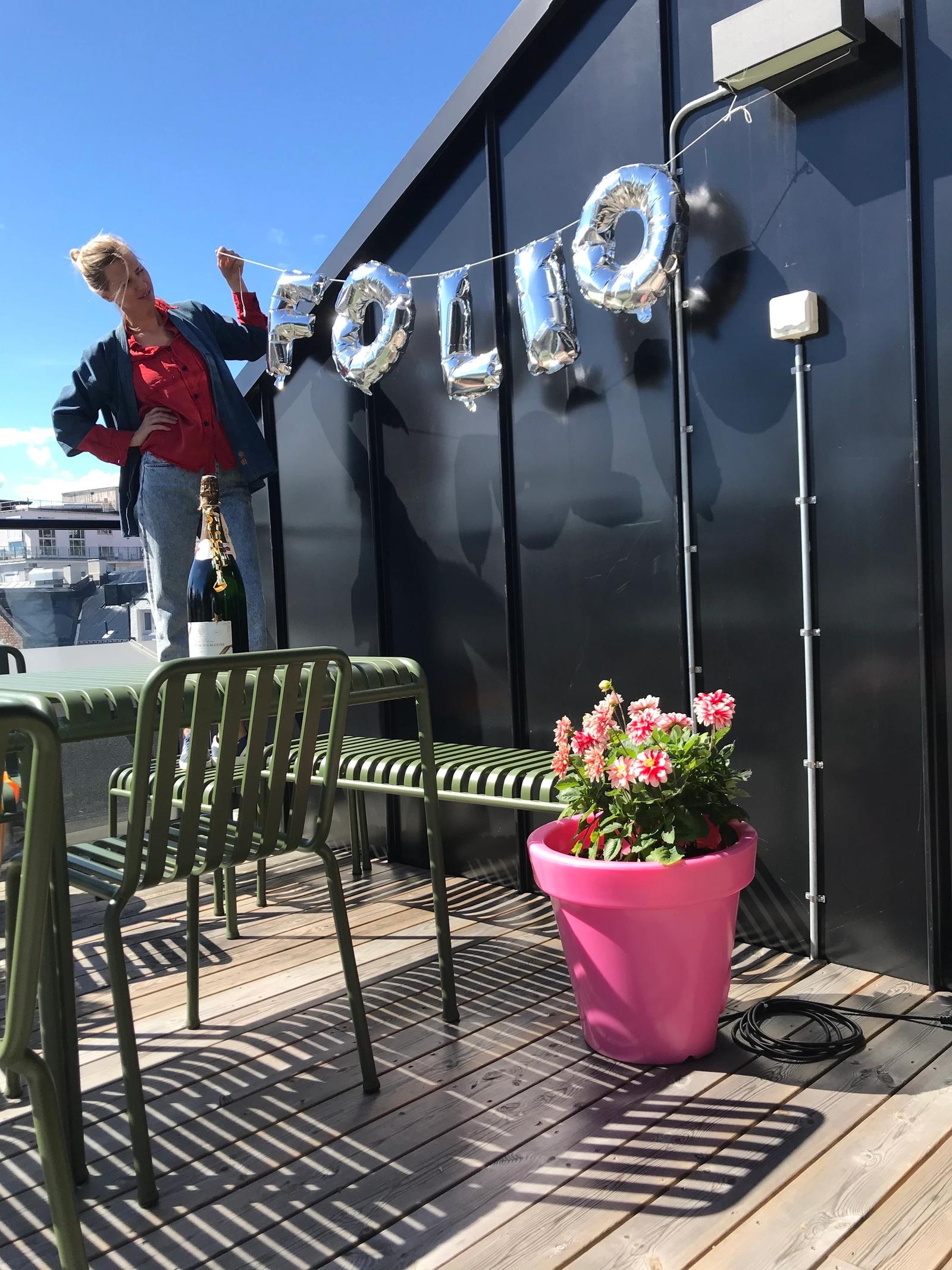 En Folio ansatt henger opp sølv-bokstavballonger som staver FOLIO på en vegg. Hun står på en takterrasse der solen skinner