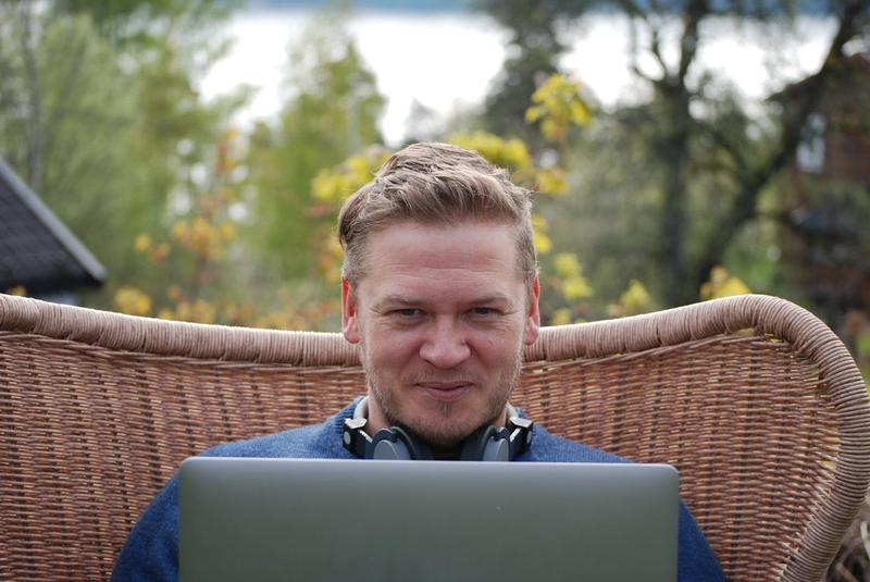 Mann sitter utendørs med laptop på fanget og smiler mot kamera.