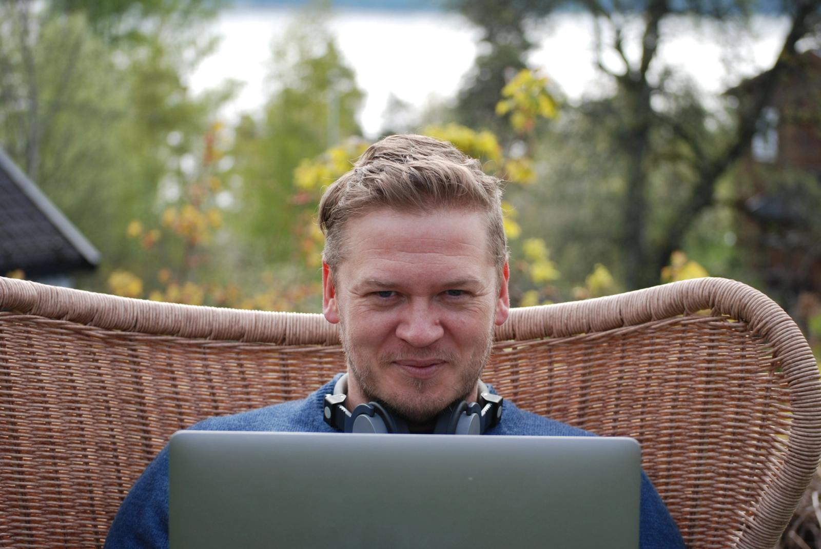 Mann sitter utendørs med laptop på fanget og smiler mot kamera.