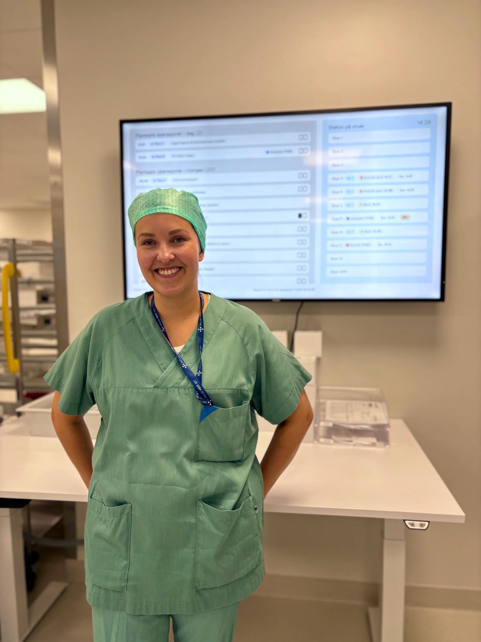 Dame i grønn sykehusuniform står framfor skjerm med brukergrensesnitt på sykehus.