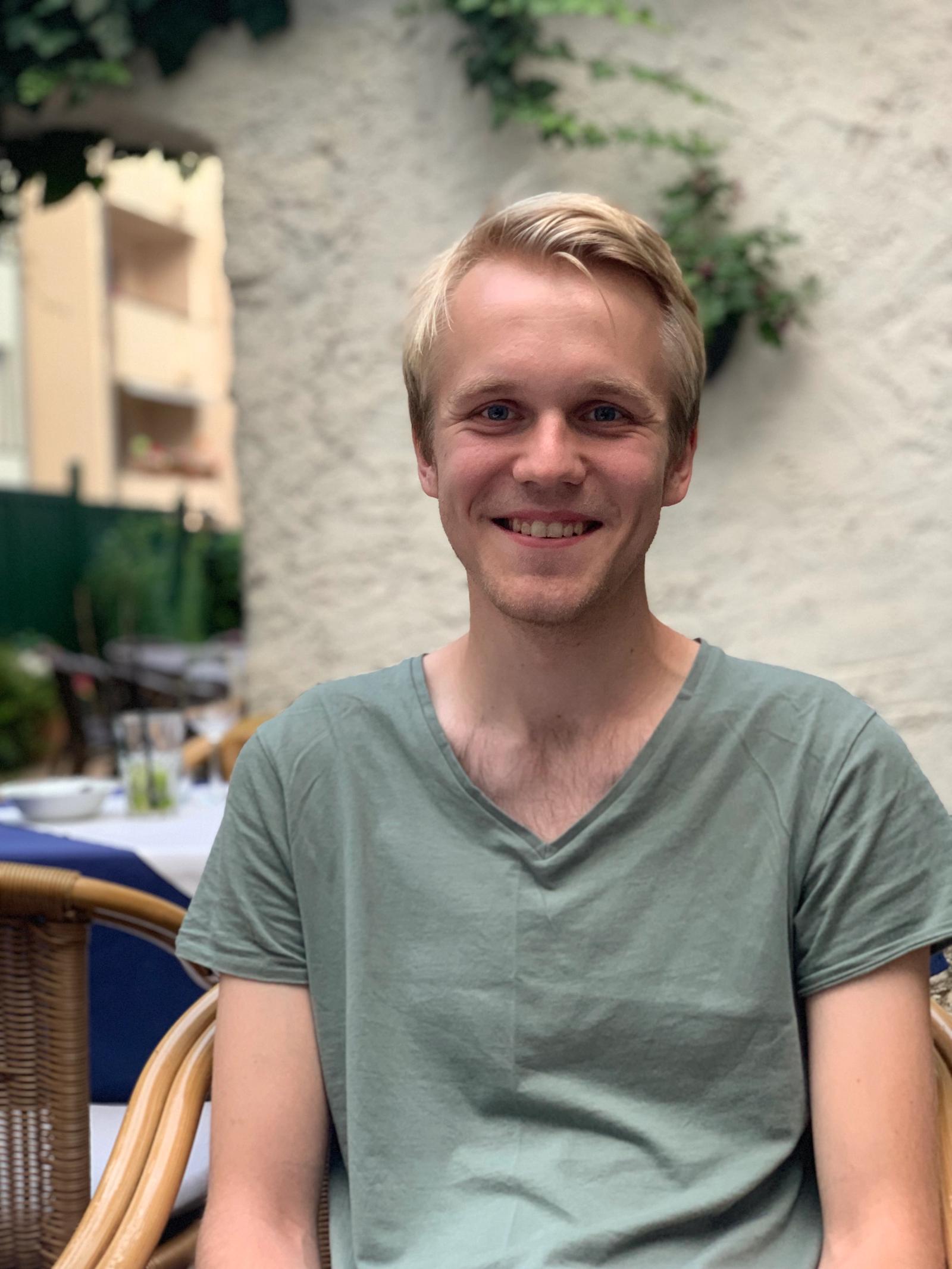 Mann med blondt hår og grønn t-skjorte sitter utendørs i sydlige strøk og smiler.