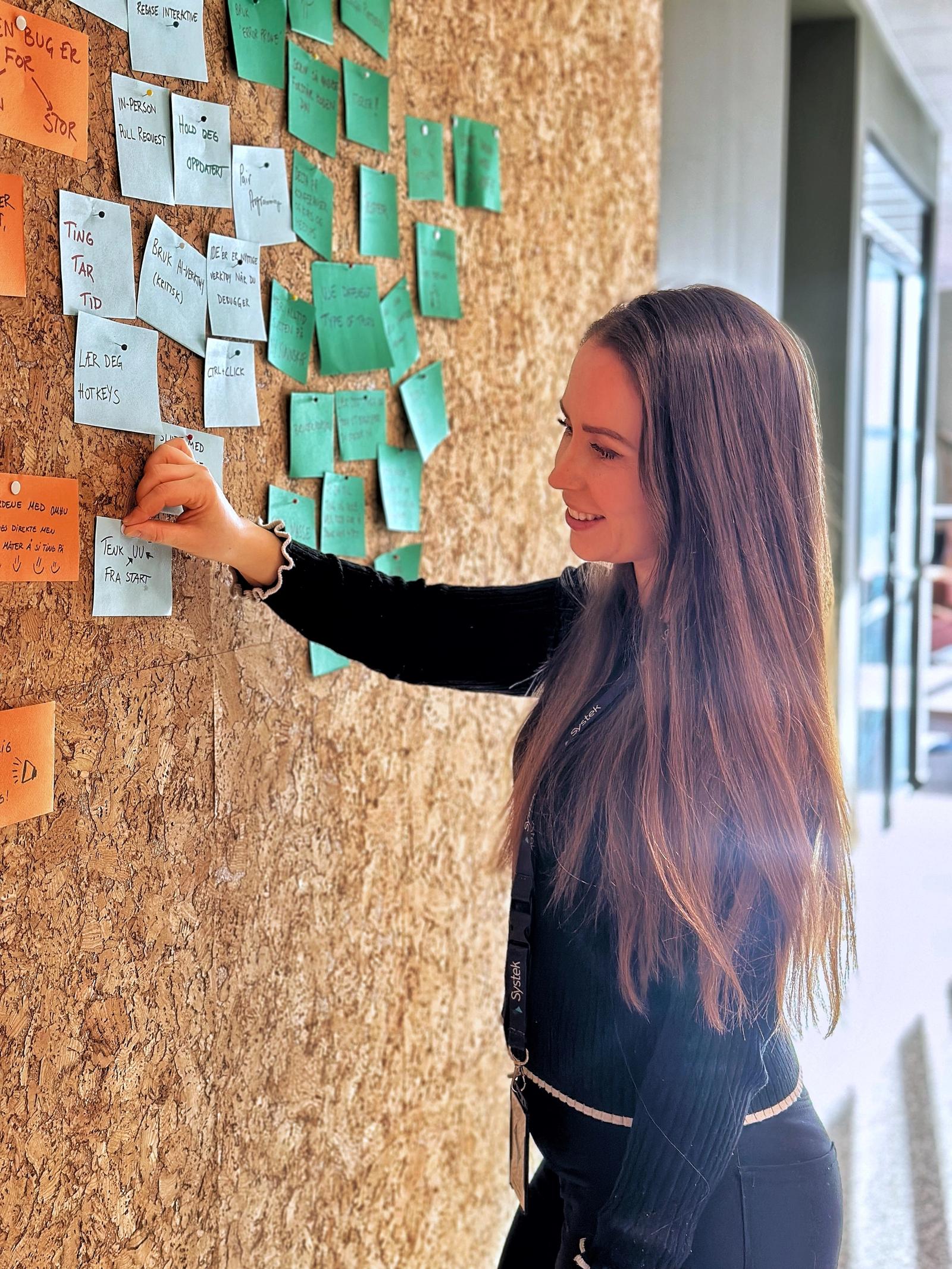 Kvinne med langt brunt hår står ved en vegg og henger opp post-it-lapper