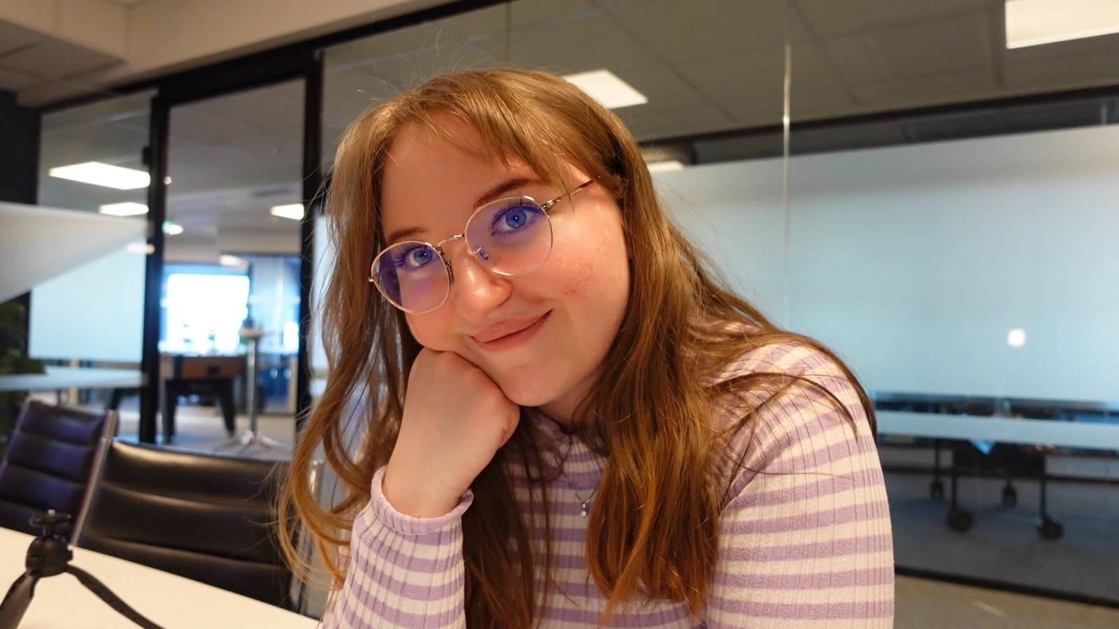 Dame med lyst, brunt hår, briller og stripete genser sitter i kontorlandskap.