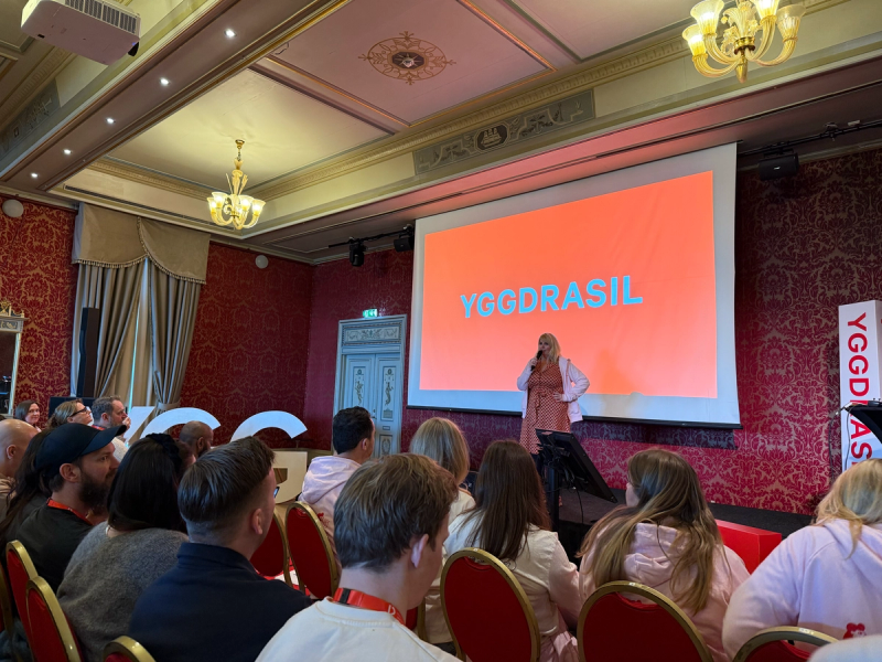 Dame står på scenen framfor publikum med teksten "yggdrasil" i store bokstaver bak seg.