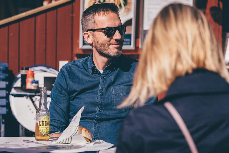 Fotoet viser Thomas med solbriller på som ser til siden. Han sitter utendørs i sollys med en burger og øl framfor seg. I forgrunnen ser man ryggen til en dame.