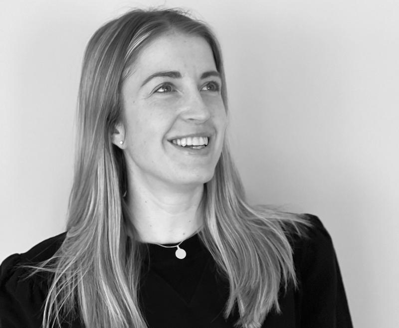 Svart hvit portrett av dame i svart genser og med smykke. Hun har langt lyst hår, smiler og ser opp og vekk fra kamera.