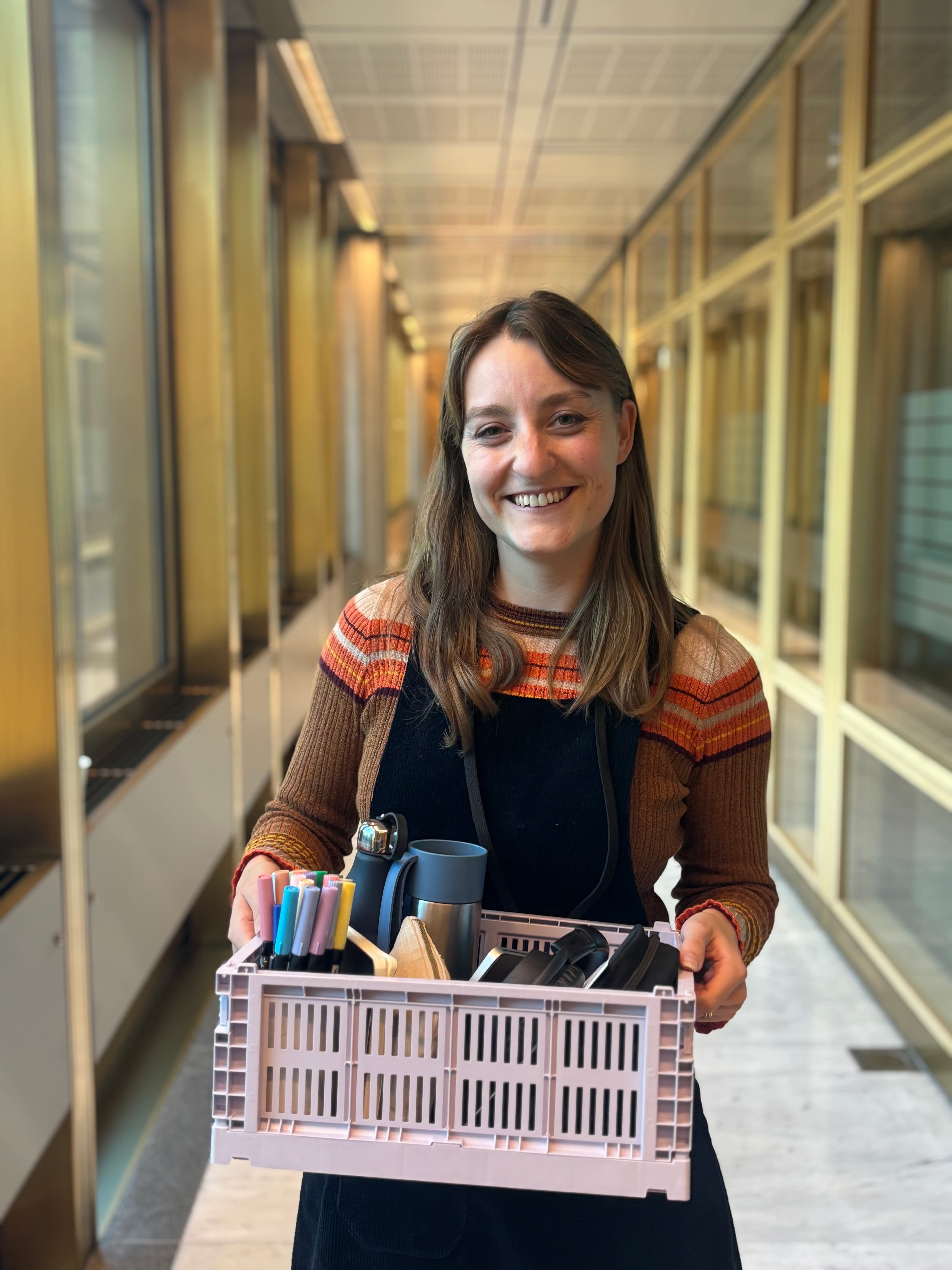 Kvinne står i en gang og holder en kurv med tusjer og termokopp i