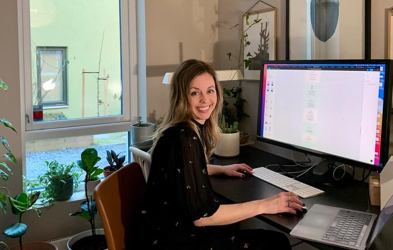 Dame sitter på kontor med flere skjermer og ser inn i kamera. Rundt henne er det mange planter og bilder på veggen.