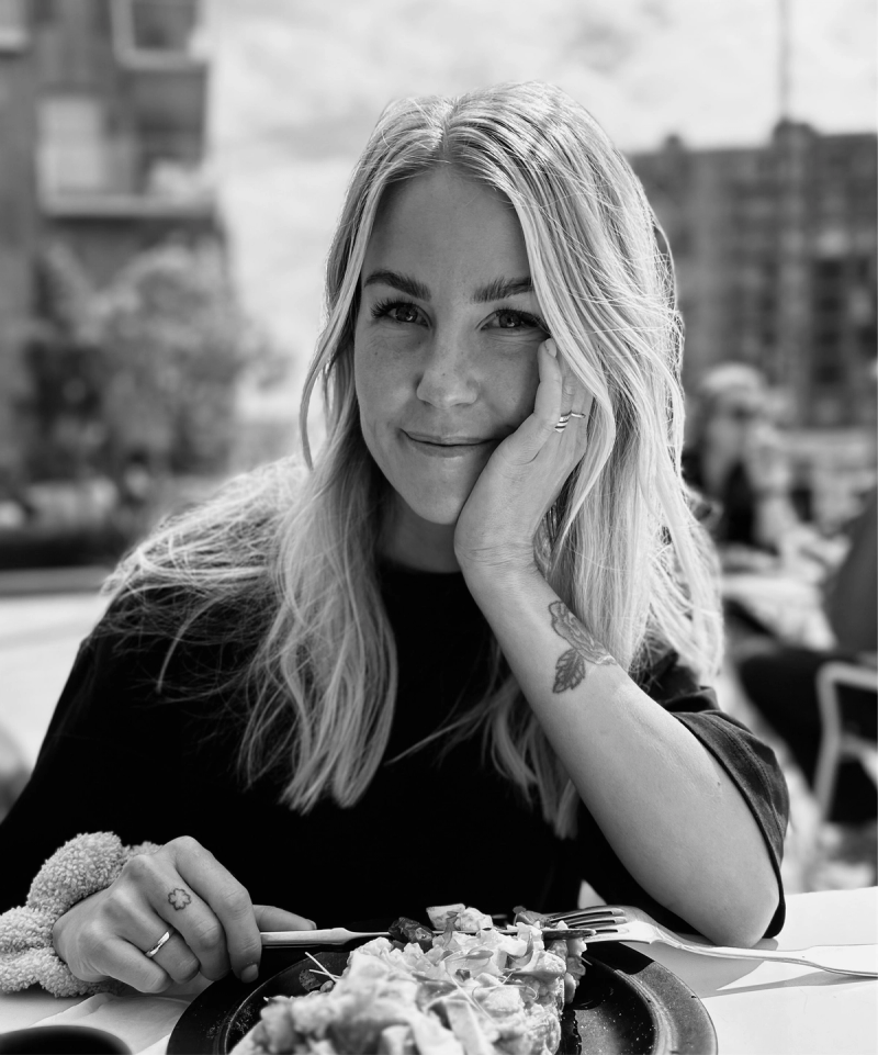 Svart-hvitt portrettfoto av dame med langt, blondt hår som sitter utendørs i bybildet.