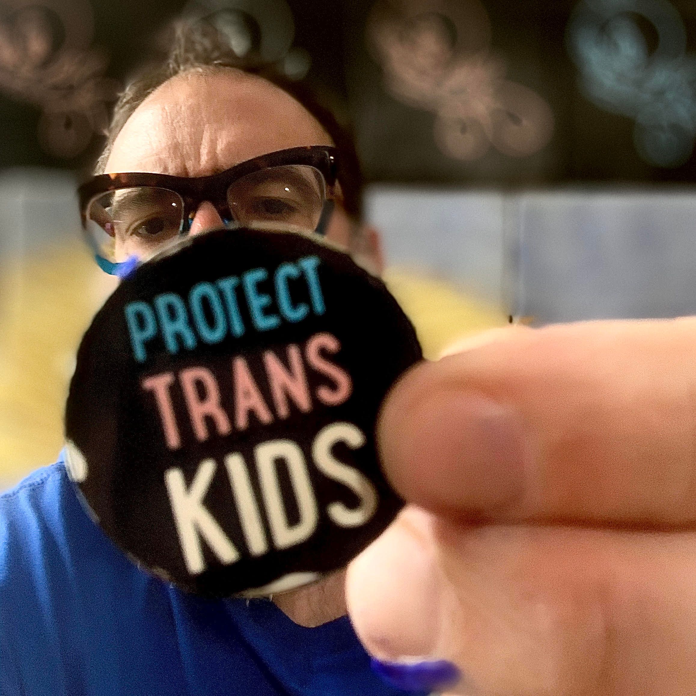 Mann med mørkt hår og briller holder et klistremerke hvor det står "Protect trans kids"