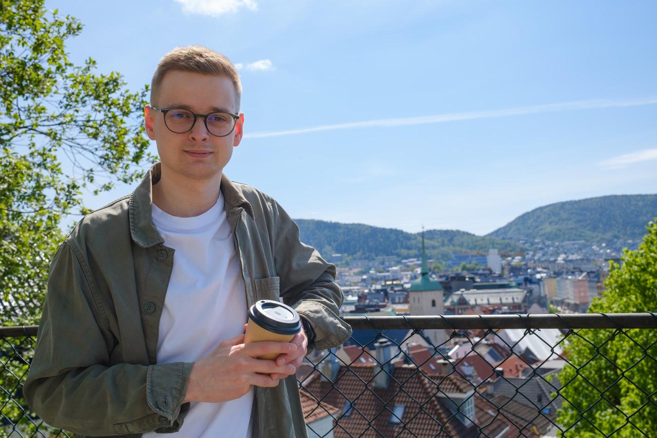 Mann poserer med kaffe i hendene i solskinn med Bergen i bakgrunnen.