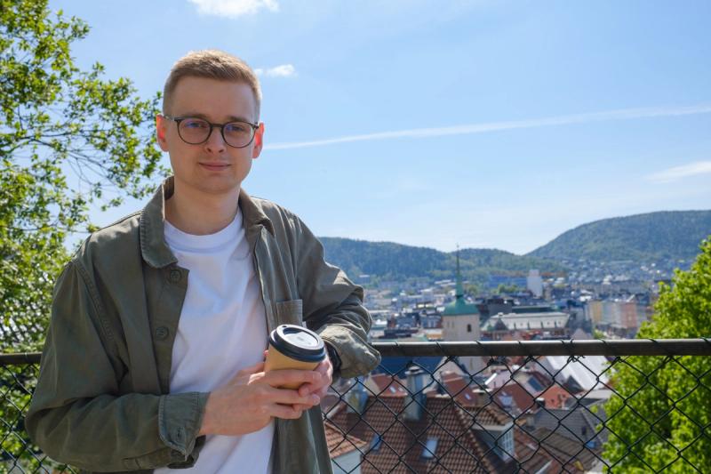 Mann poserer med kaffe i hendene i solskinn med Bergen i bakgrunnen.