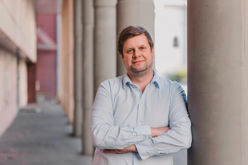 Mann i skjorte står utendørs og lener seg inntil en stor stolpe.