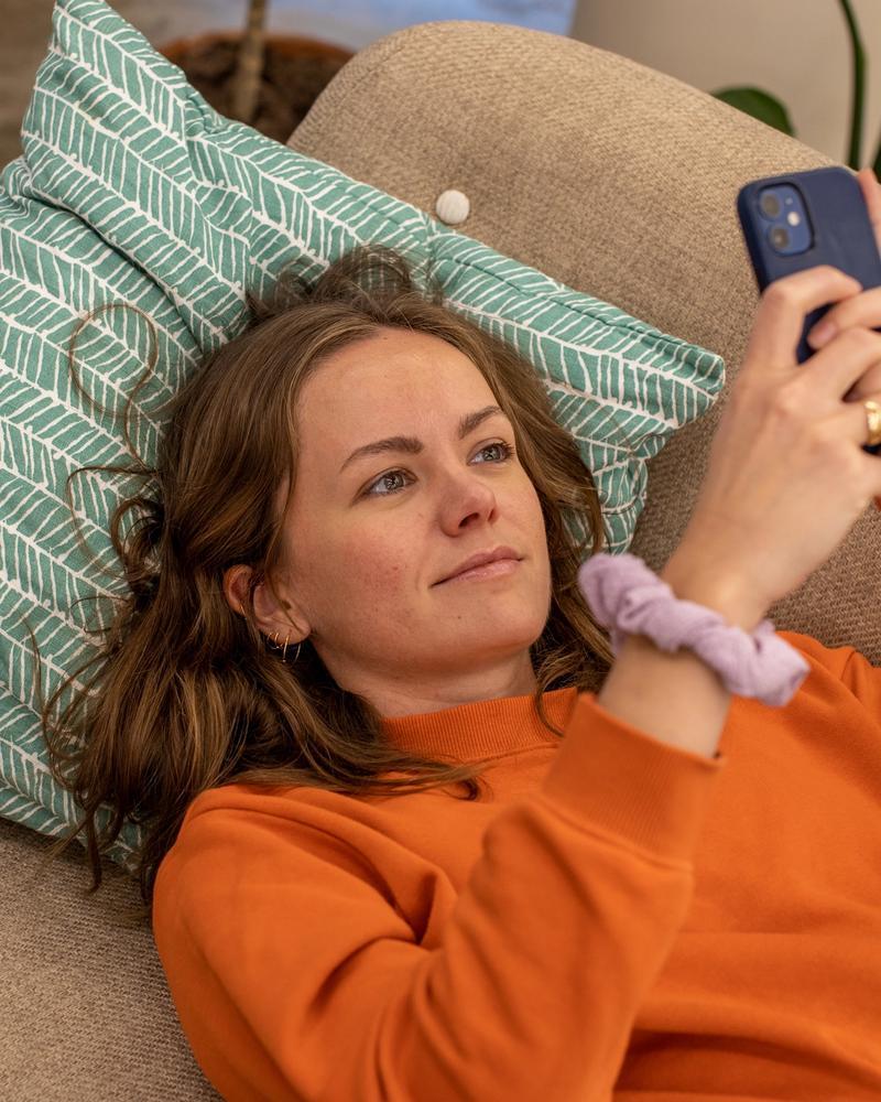 Dame med orange genser ligger i en sofa og scroller på mobilen.